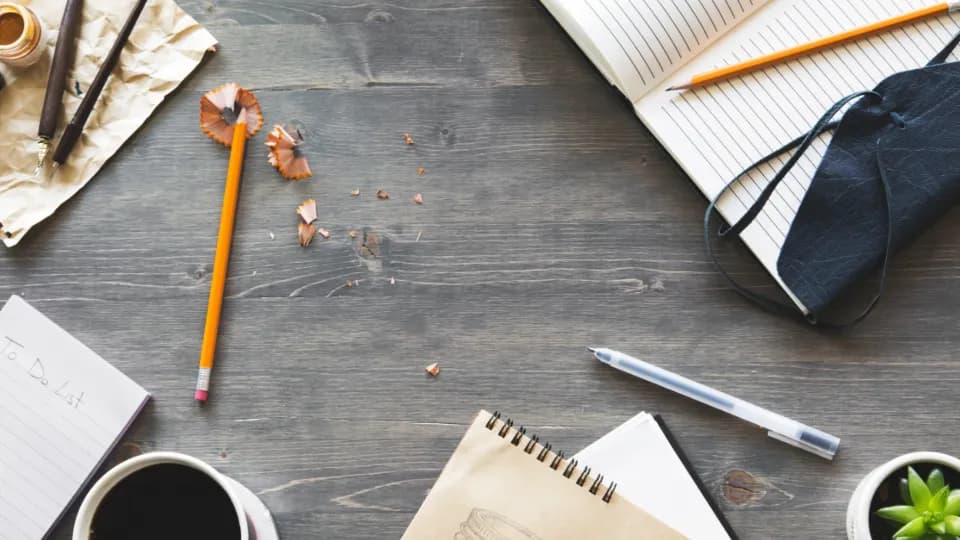 A notebook, pencils, a cup of coffee and a small plant laid out on a wooden desk