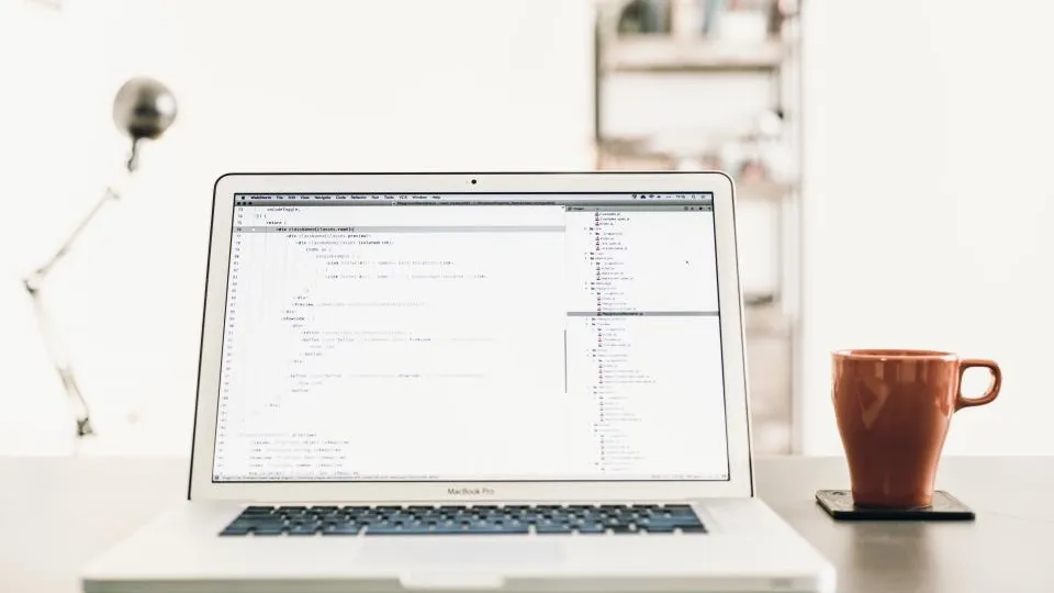 A laptop showing code on a bright white desk next to an orange coffee mug