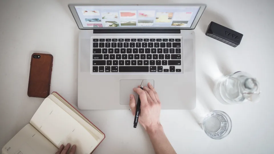 Overhead view of a hand holding a pen over a laptop, with an open notebook and a phone on a white desk